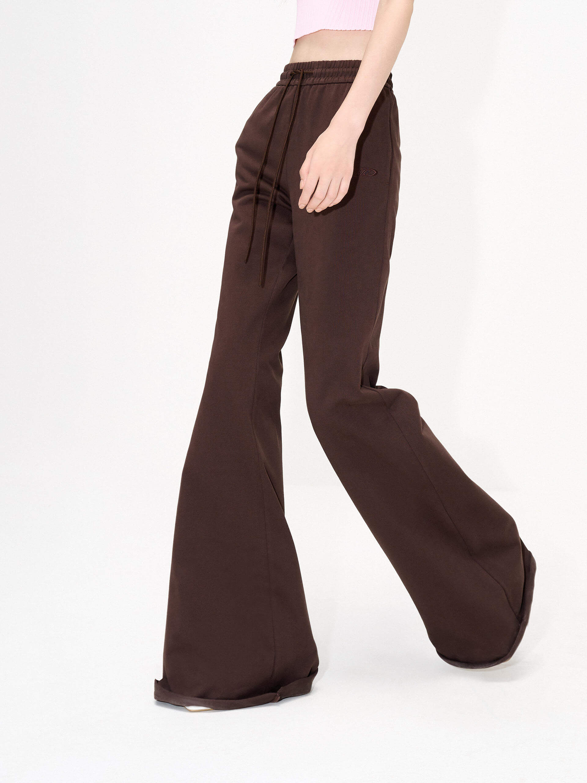 MO&Co.'s Drawstring Waist Causal Flared Sweatpants in Brown. Crafted from soft cotton, these trousers feature a relaxed fit with drawstrings and elastic waistband for comfort. They also come with flared legs and double side pockets with MC embroidery details.