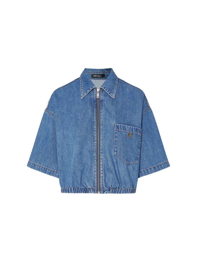 MO&Co.'s Women's Cropped Denim Jacket featuring an elasticized hem design, front zipper closure, wide and short sleeves, and front chest pocket with metallic logo.