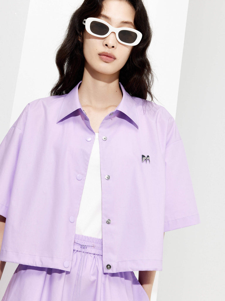 MO&Co. Women's Boxy Fit Cotton Shirt Jacket in Short Sleeves in purple features cropped, short sleeves, a button closure, and a M metallic embellishment at the front. Choose from color in purple and black, and pair with the matching shorts for a stylish look.